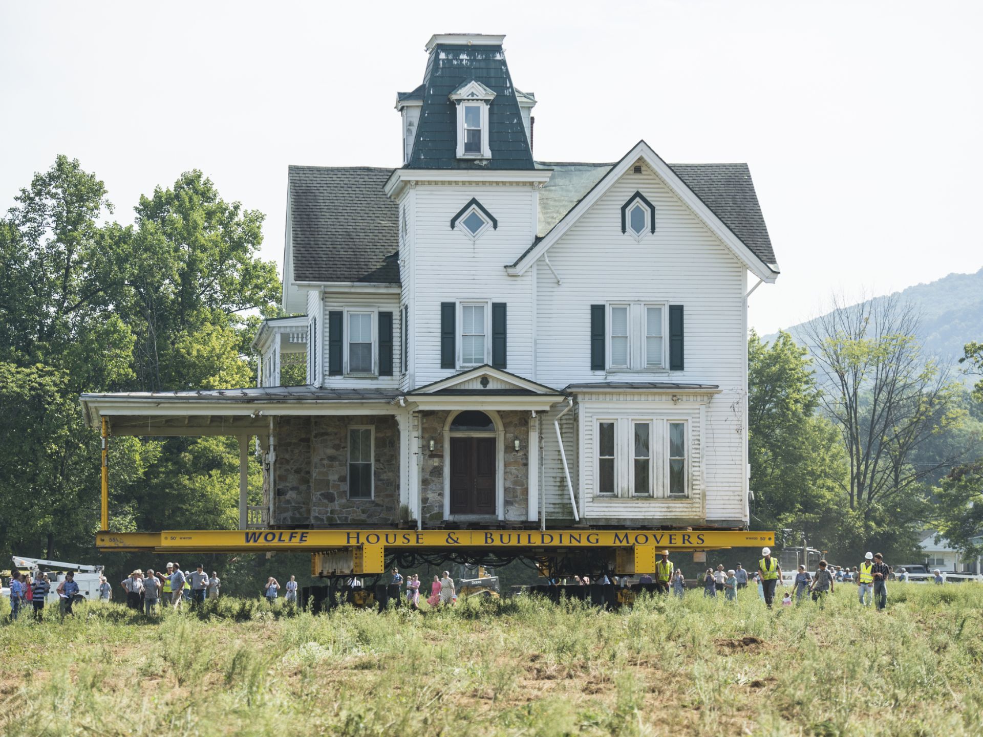 Newman house raised on dolly moving through grass field as dozens of bystanders walk with it.