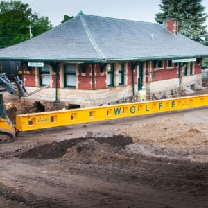 Two skid loaders hoisting up each end of steel beam next to the Sturgis train depot.