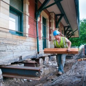 Worker inserting wooden block underneath a red brick and stone structure structure.