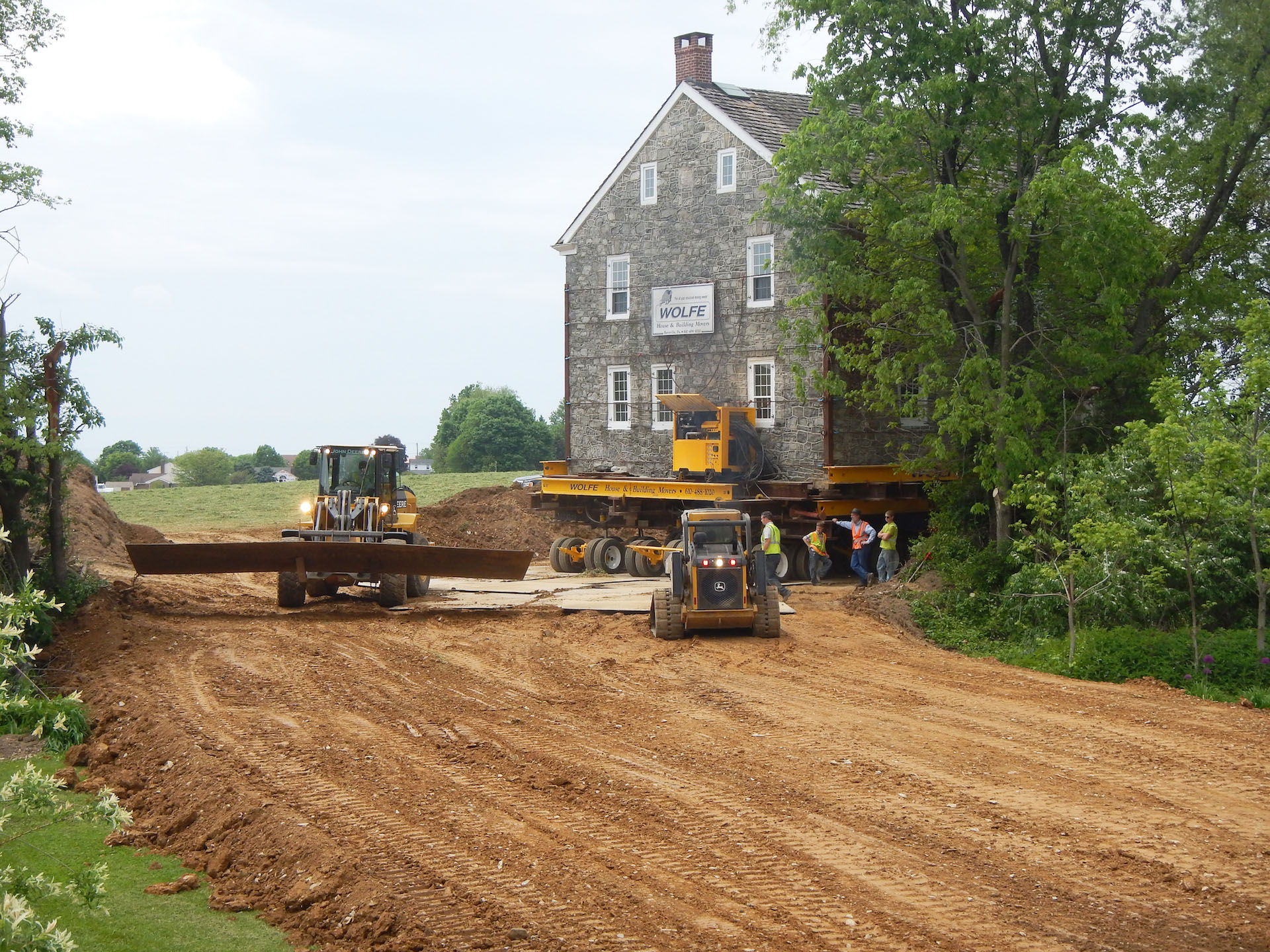 Skid loaders placing plywood in front of a building being moved on a dirt path.