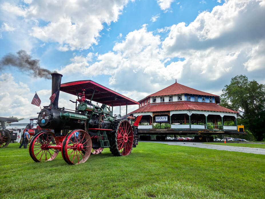 A vintage steam engine chugs smoke in the foreground with the carousel house behind it