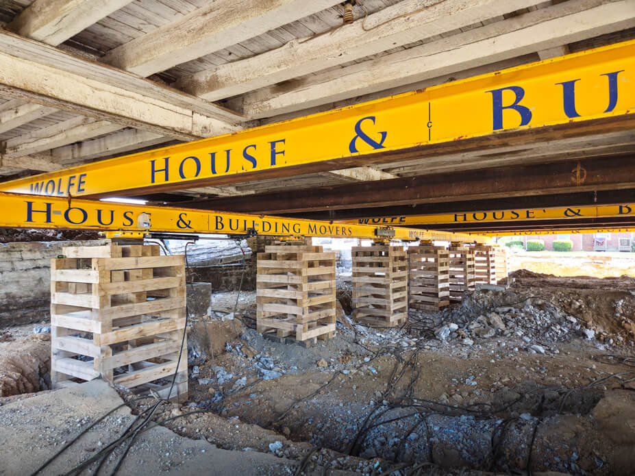 Yellow Wolfe House Movers beams support barn while push-pull rams slide the building over rollers inside crib piles